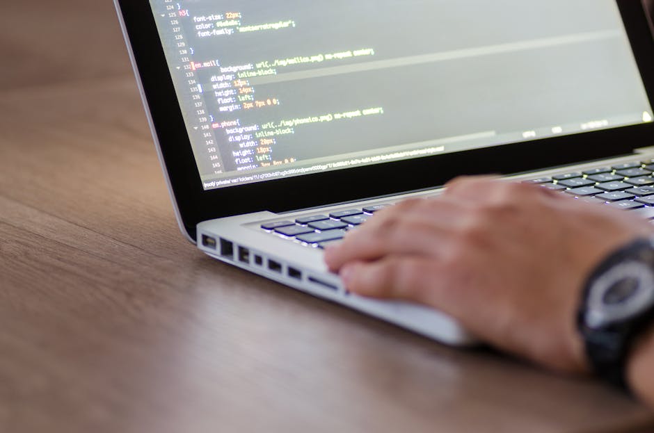 A close-up image of a person's hand operating a silver laptop on a wooden surface, with the laptop screen displaying lines of code in a text editor. The person is wearing a black wristwatch, and the e