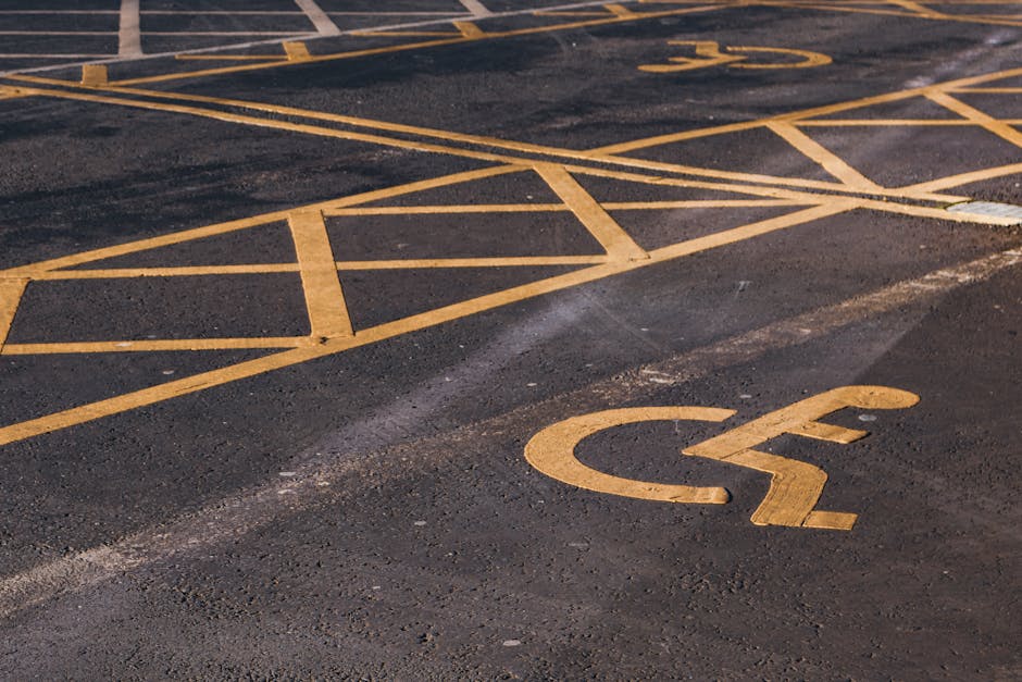 A close-up view of an outdoor parking area surface with yellow paint markings, including a designated disabled parking space identified by the wheelchair symbol. The asphalt ground appears dark and te